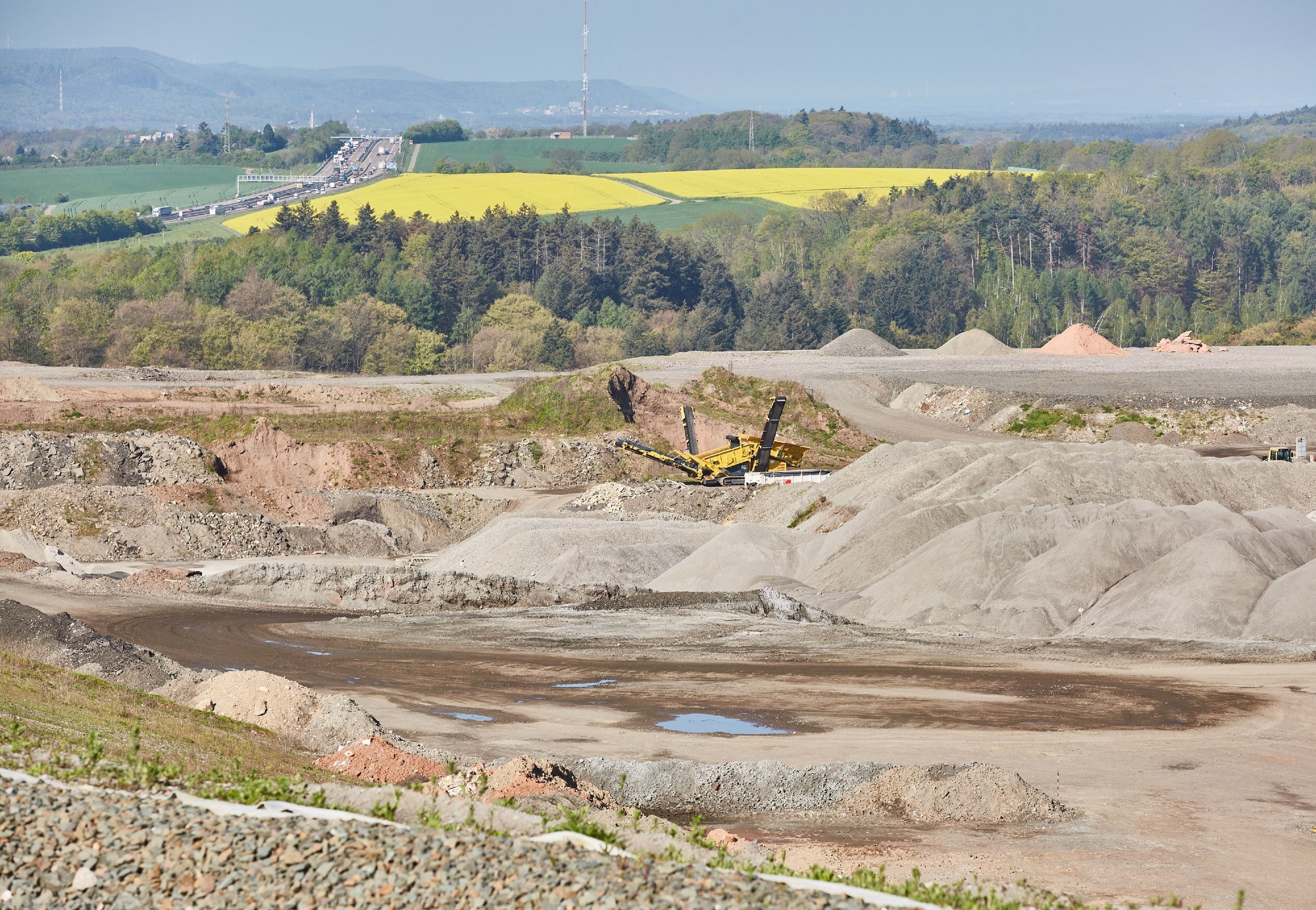 ZAK Deponie-Bauarbeiten mit Bagger in weitläufiger Grube vor Kaiserslauterer Landschaftskulisse mit Rapsfeldern