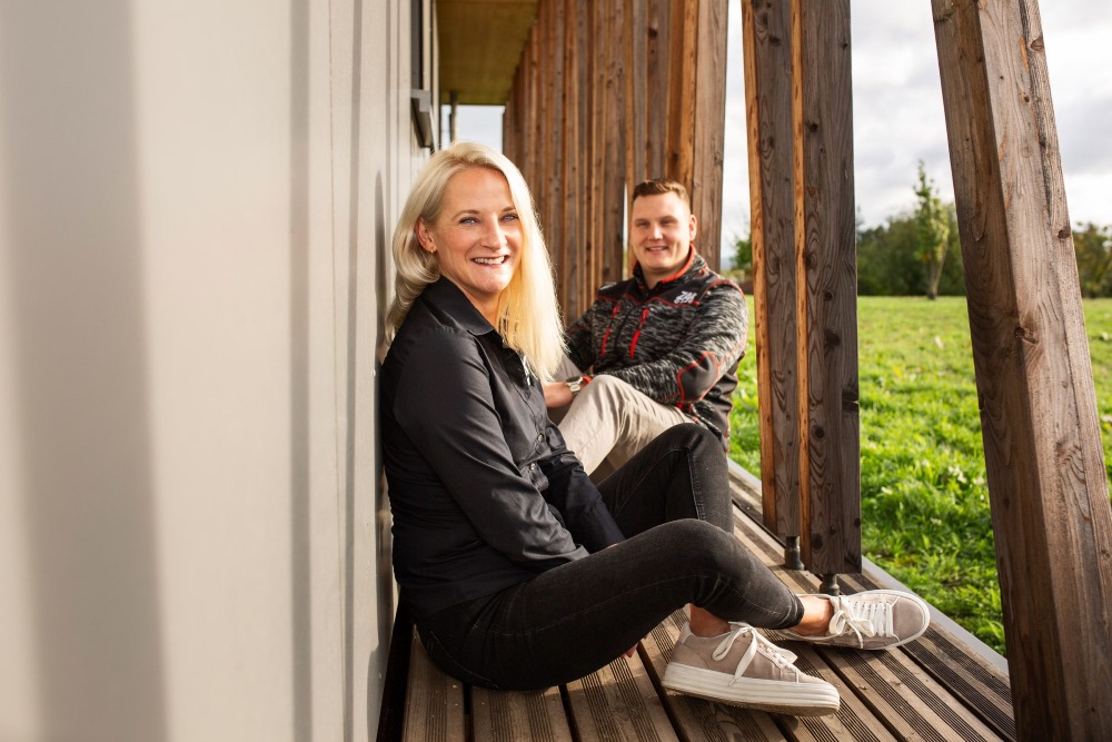 Zwei Mitarbeitende sitzen vor dem Gebäude Haus Aspenkopf und lachen in die Kamera.